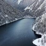 Winter view of Kolsay Lake, Almaty Region, Kazakhstan, with snow-covered Tien Shan mountains, evergreen forest, and calm blue water at sunrise.